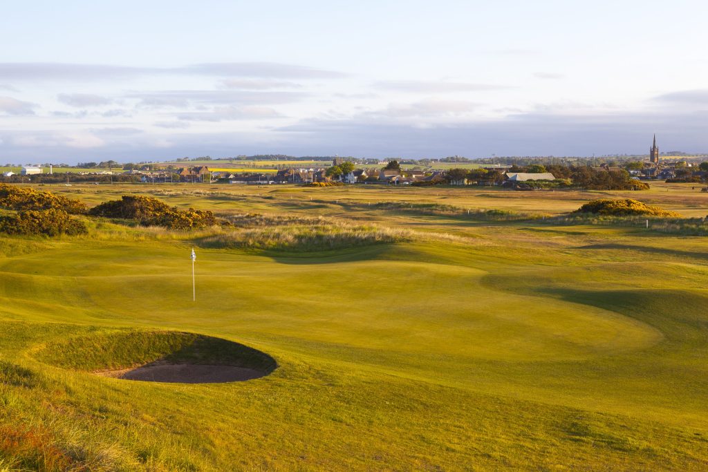 A golf course with a green, a flagstick, and a sand bunker in the foreground. The landscape is open and grassy, with small buildings and a church spire visible in the distance under a partly cloudy sky.