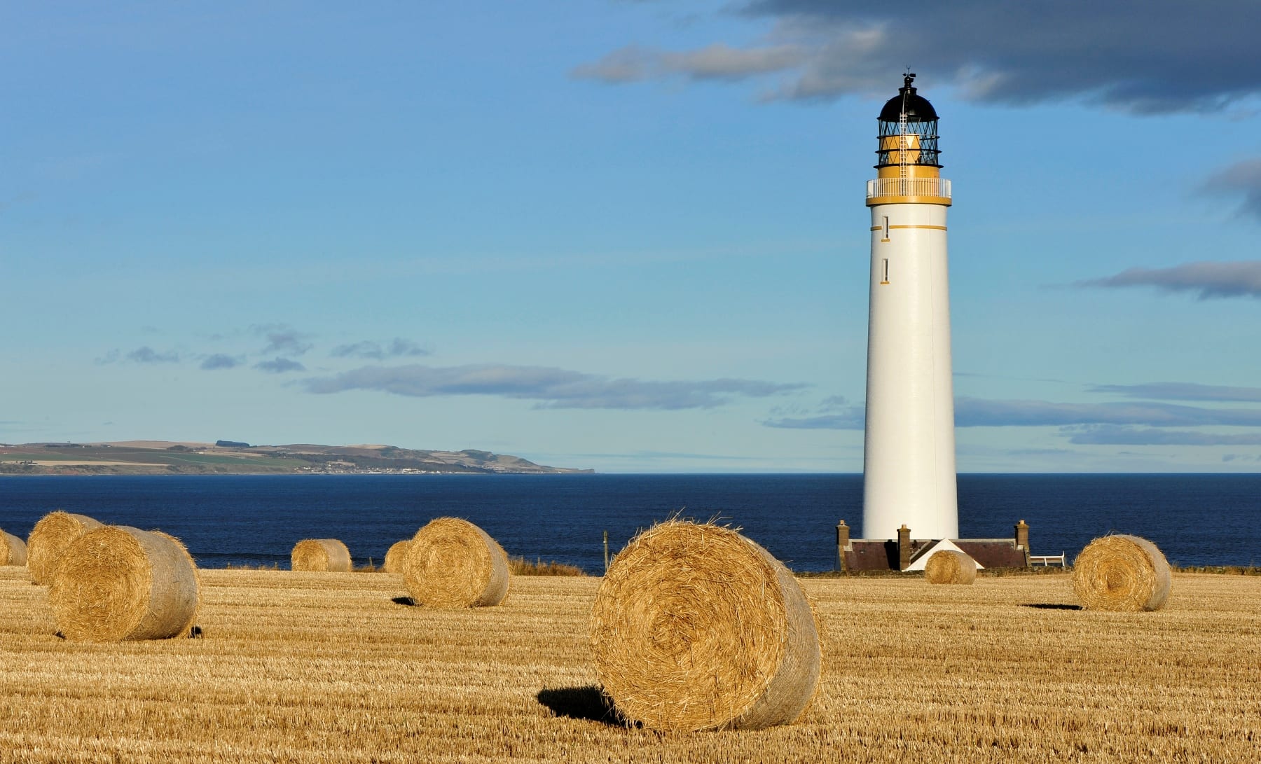 A tall white lighthouse with a black top stands near the sea, surrounded by a field with several large round hay bales under a partly cloudy sky.