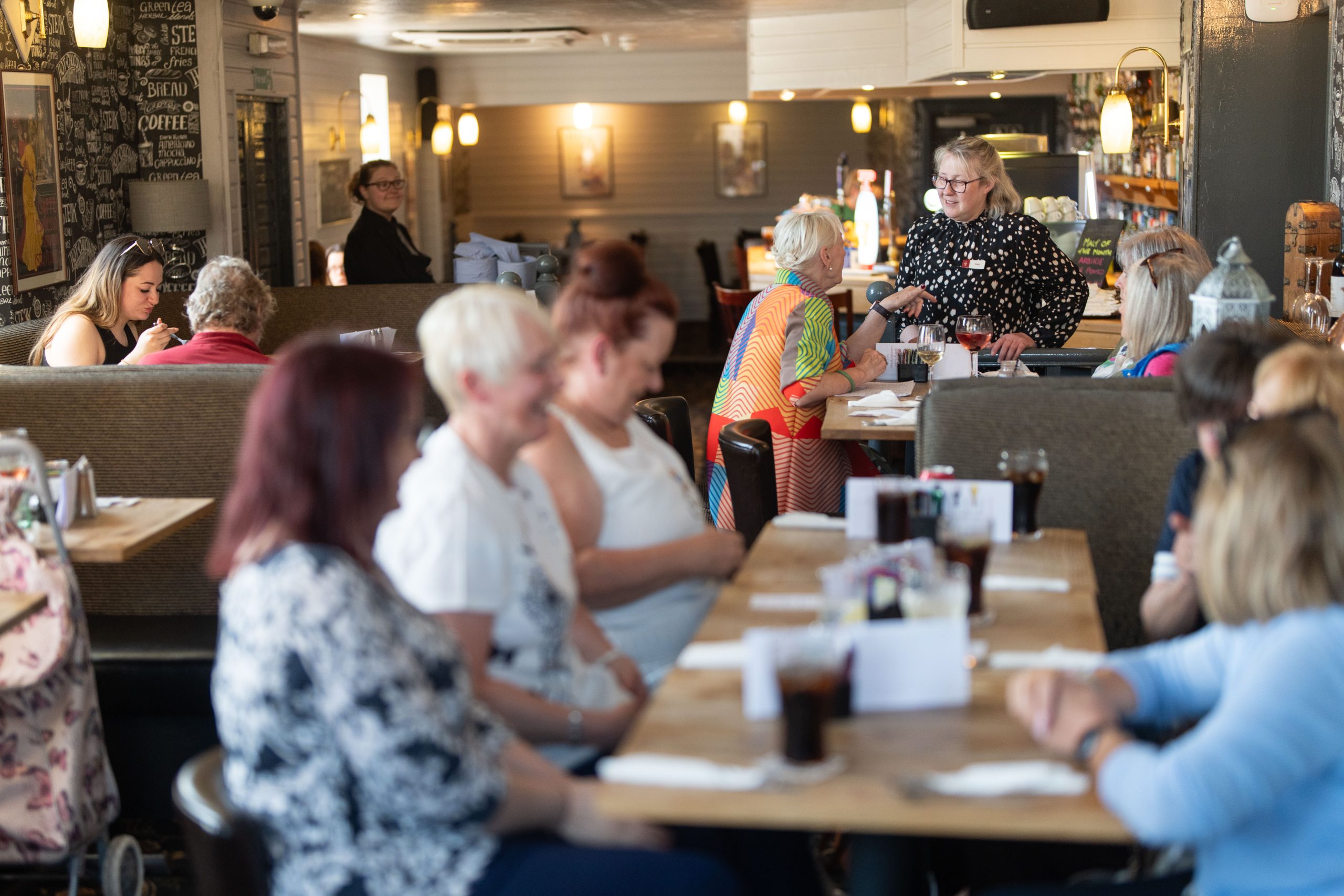 A group of women sit at a long table in a busy café or restaurant, talking and drinking beverages. Other patrons are seated at separate tables in the background. The atmosphere is casual and lively.
