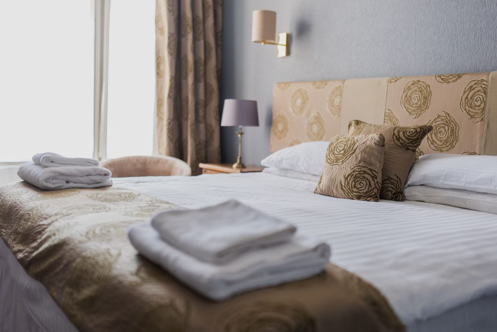 A neatly made bed with folded white towels, patterned beige bedding, pillows, a bedside table with a lamp, and a window with curtains letting in natural light.