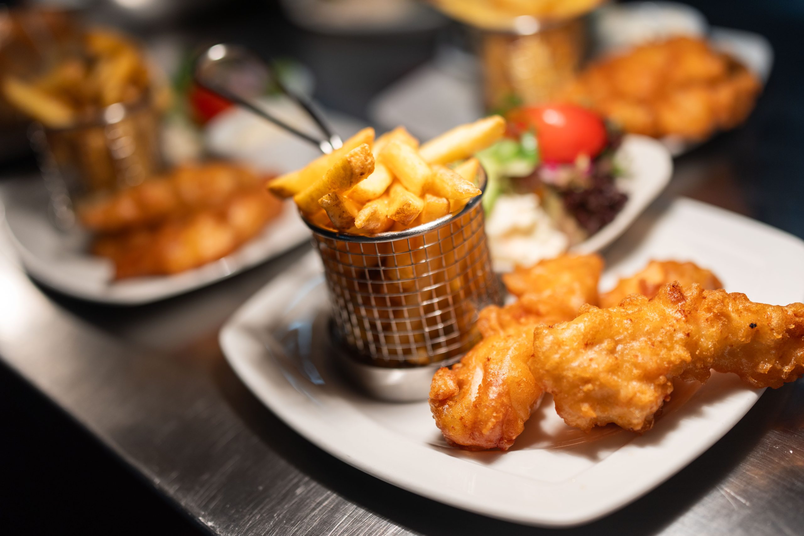 A plate with battered fried fish and golden chips served in a small metal basket, with a side salad and dipping sauce in the background, on a white square plate.