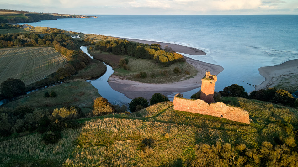 A drone view of a ruined stone tower on a grassy hill overlooking a winding river, fields, trees, and a sandy coastline, with the sea and sky in the background.
