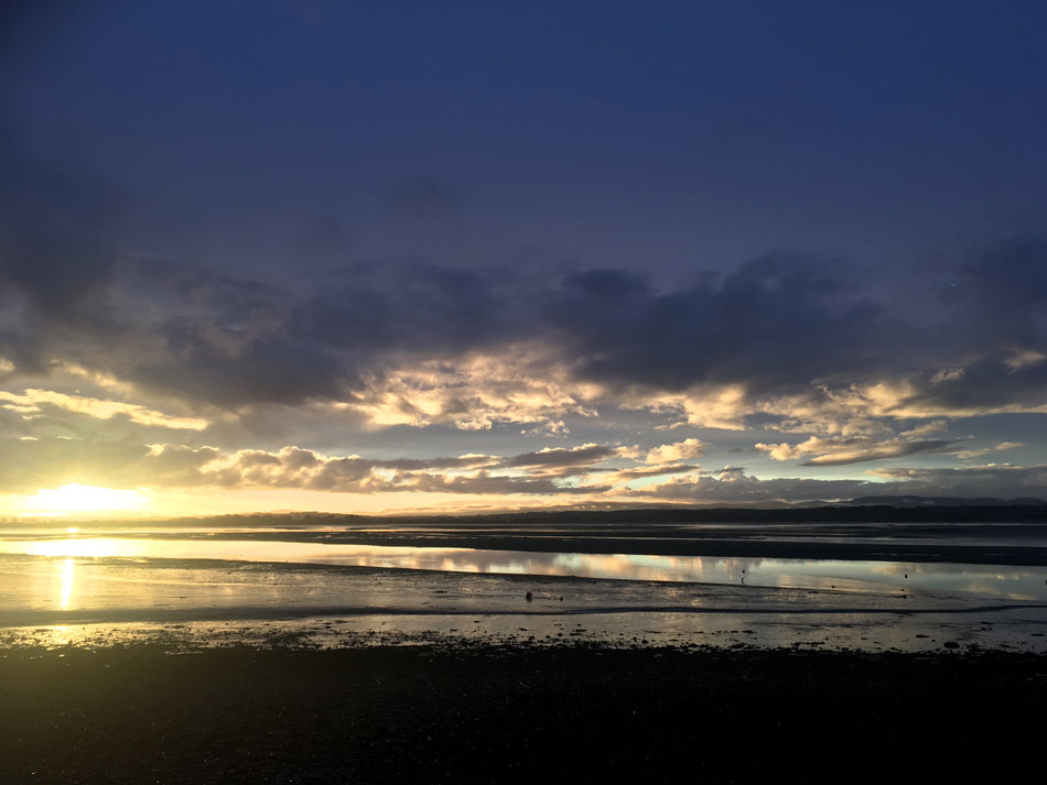 A calm beach scene at sunset with a wide sky filled with clouds, the sun low on the horizon, and reflections of the sky visible on the wet sand and shallow water.