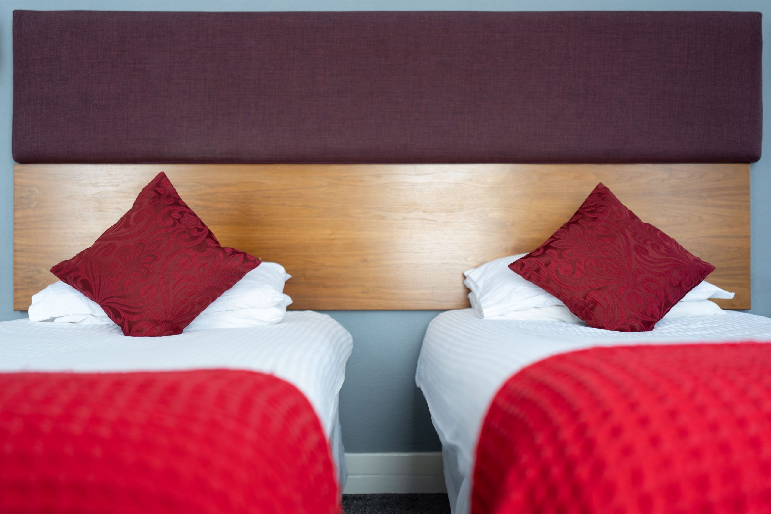 Two single beds with white linen and red textured blankets are side by side. Each bed has a red decorative cushion resting against a stack of white pillows. A wooden headboard with a dark upper section is behind the beds.