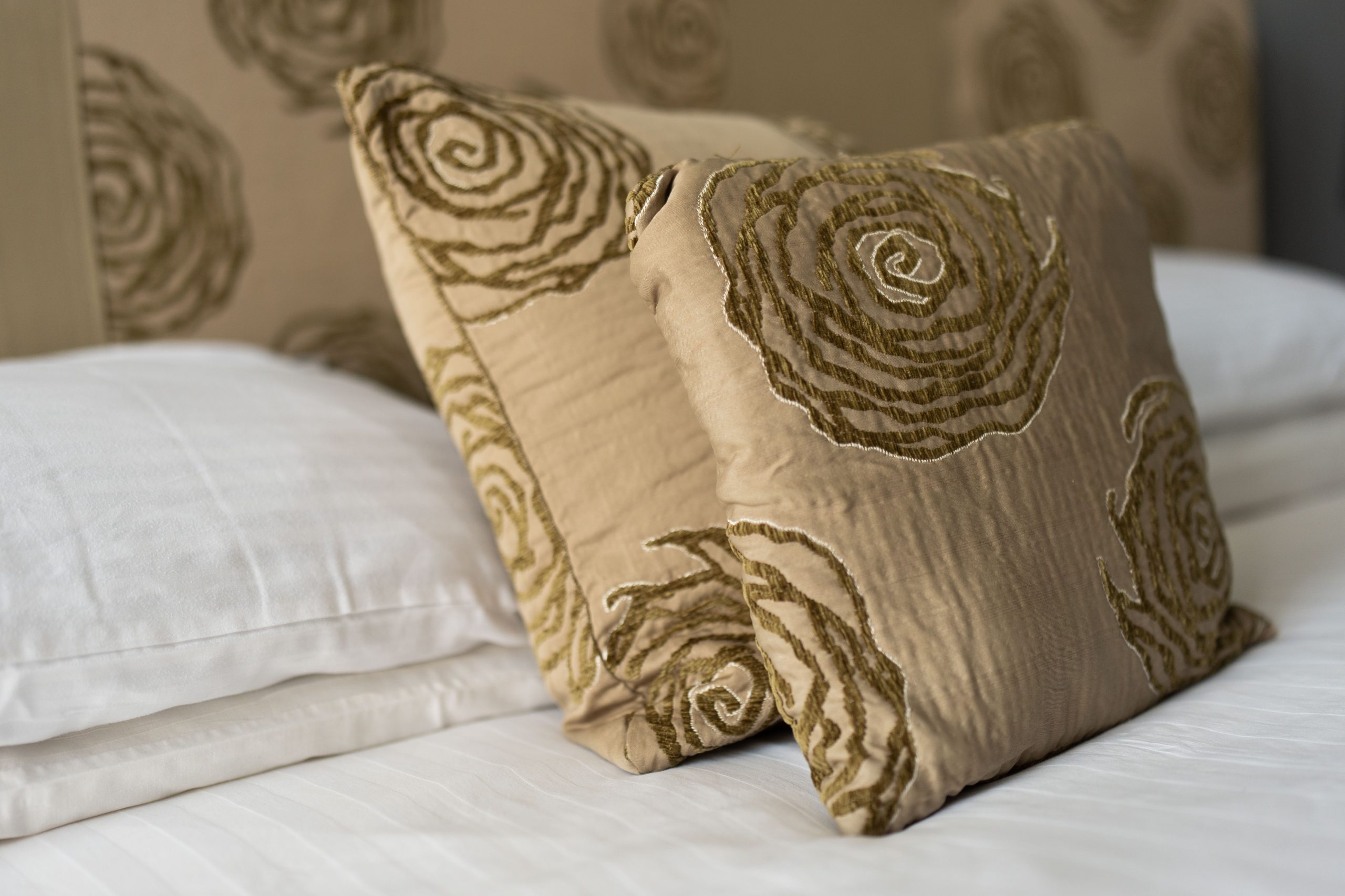 Two decorative cushions with a textured, spiral pattern in beige and green rest against white pillows on a neatly made bed with white sheets. The headboard in the background has a matching spiral design.