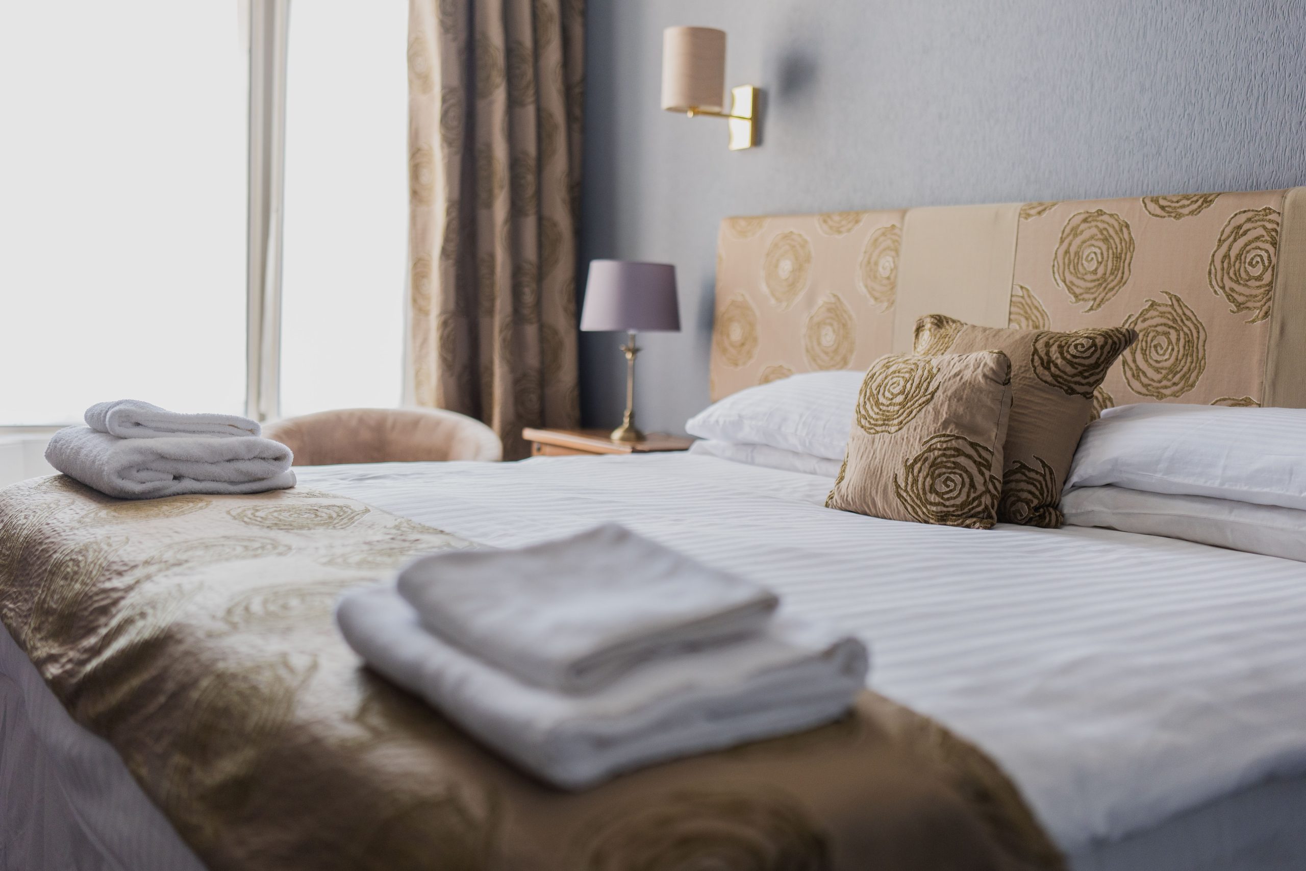 A neatly made bed with patterned gold and beige bedlinen and pillows, folded white towels at the foot, a bedside lamp, and a window with curtains in a softly lit hotel room.