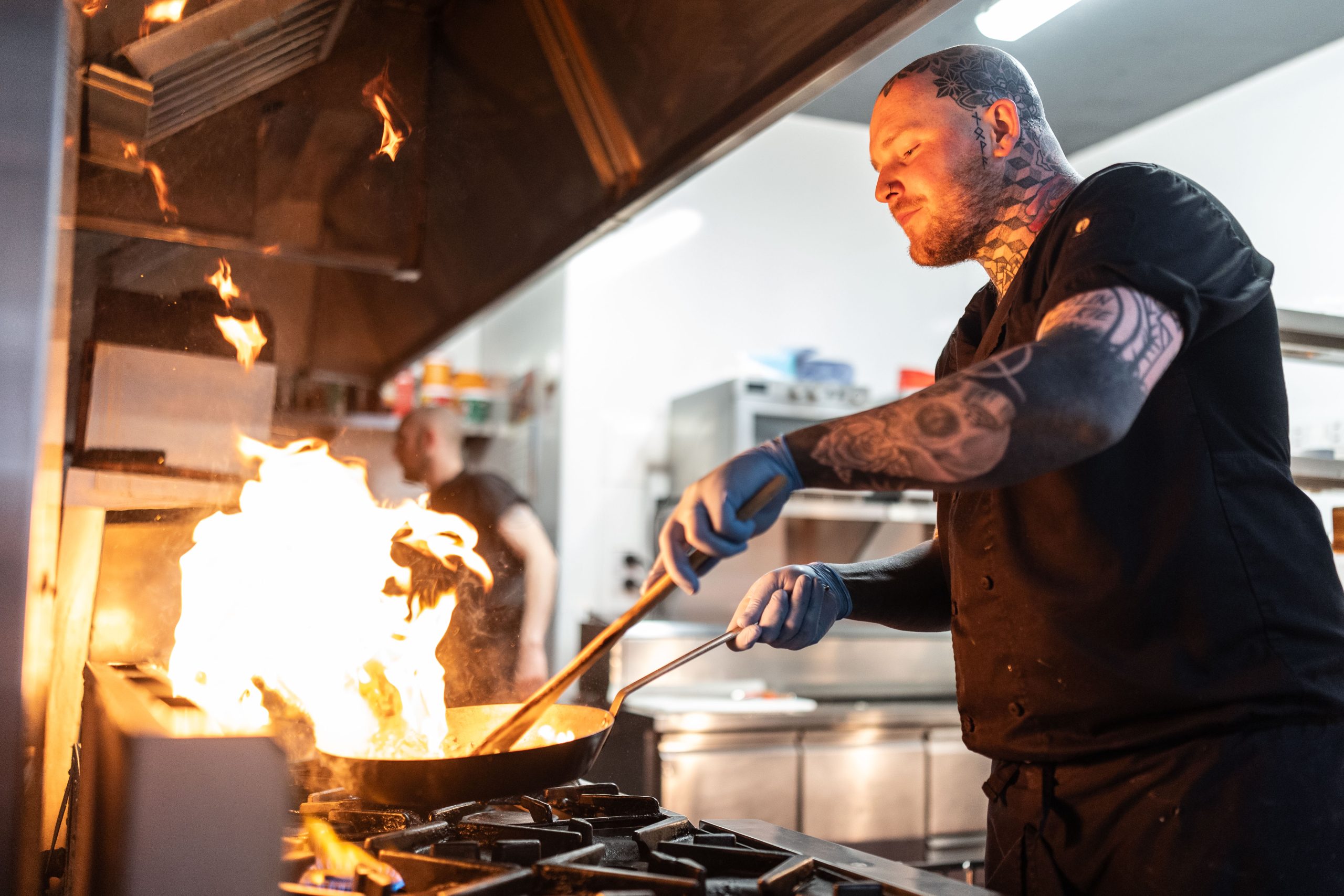 A chef with tattoos and gloves is cooking at a cooker, holding a pan with visible flames rising from it in a commercial kitchen.
