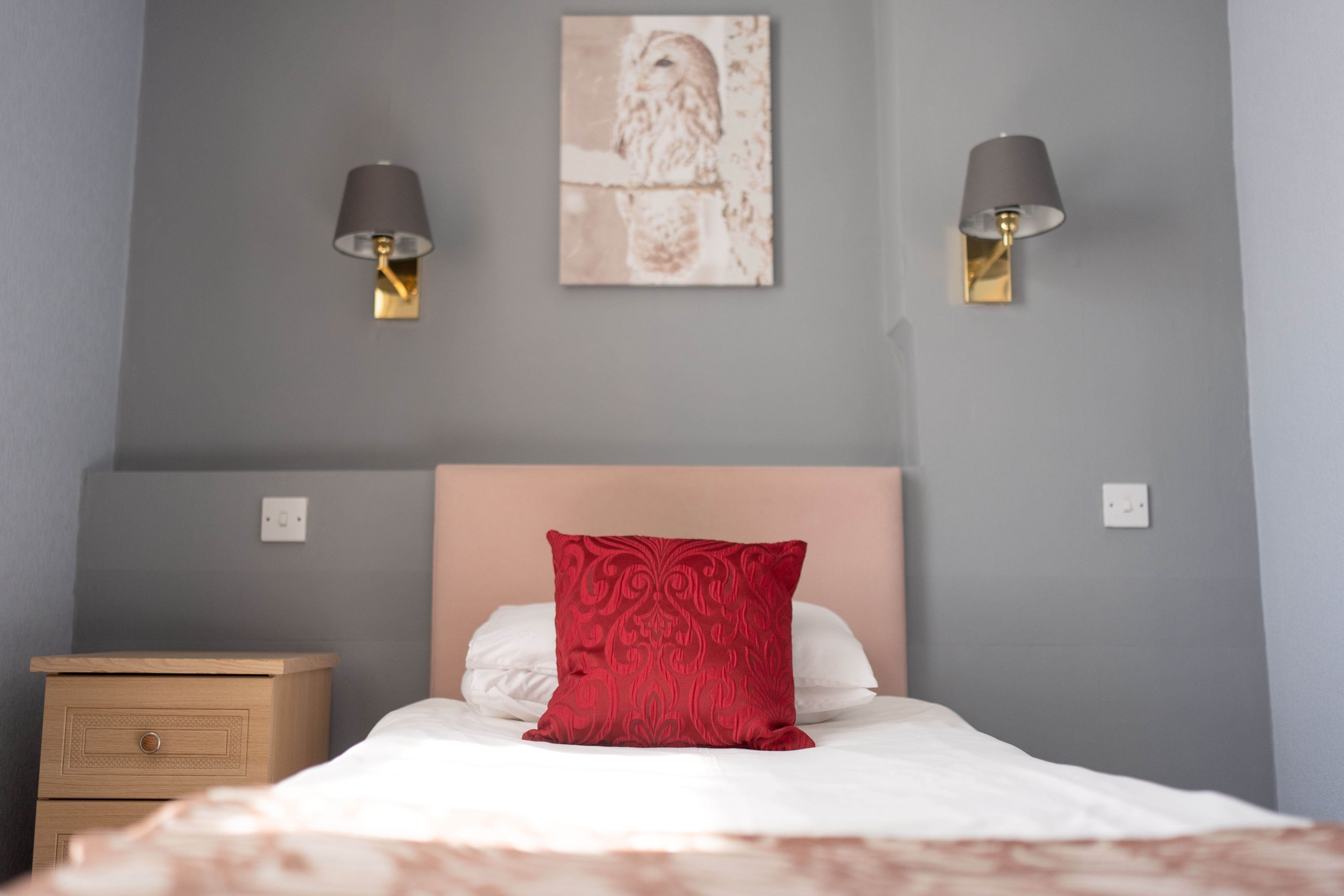 A neatly made single bed with a red decorative cushion, a light-coloured headboard, and a wooden bedside table. Two wall lamps and a framed owl artwork hang above the bed, against a grey wall.