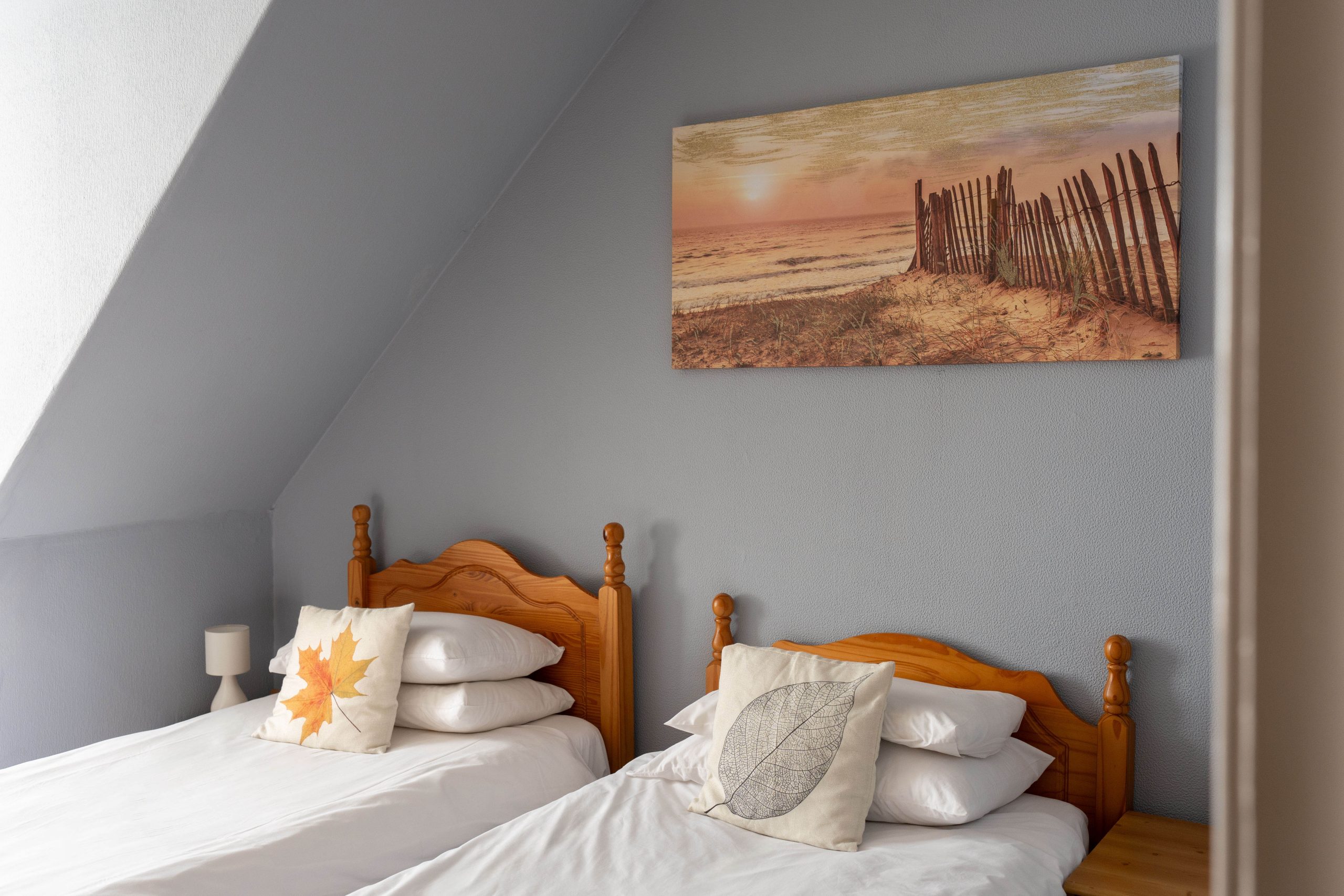 Two single beds with wooden headboards are placed side by side against a grey wall. Each bed has white linen and a decorative cushion. Above the beds hangs a landscape painting of a beach scene with a fence.