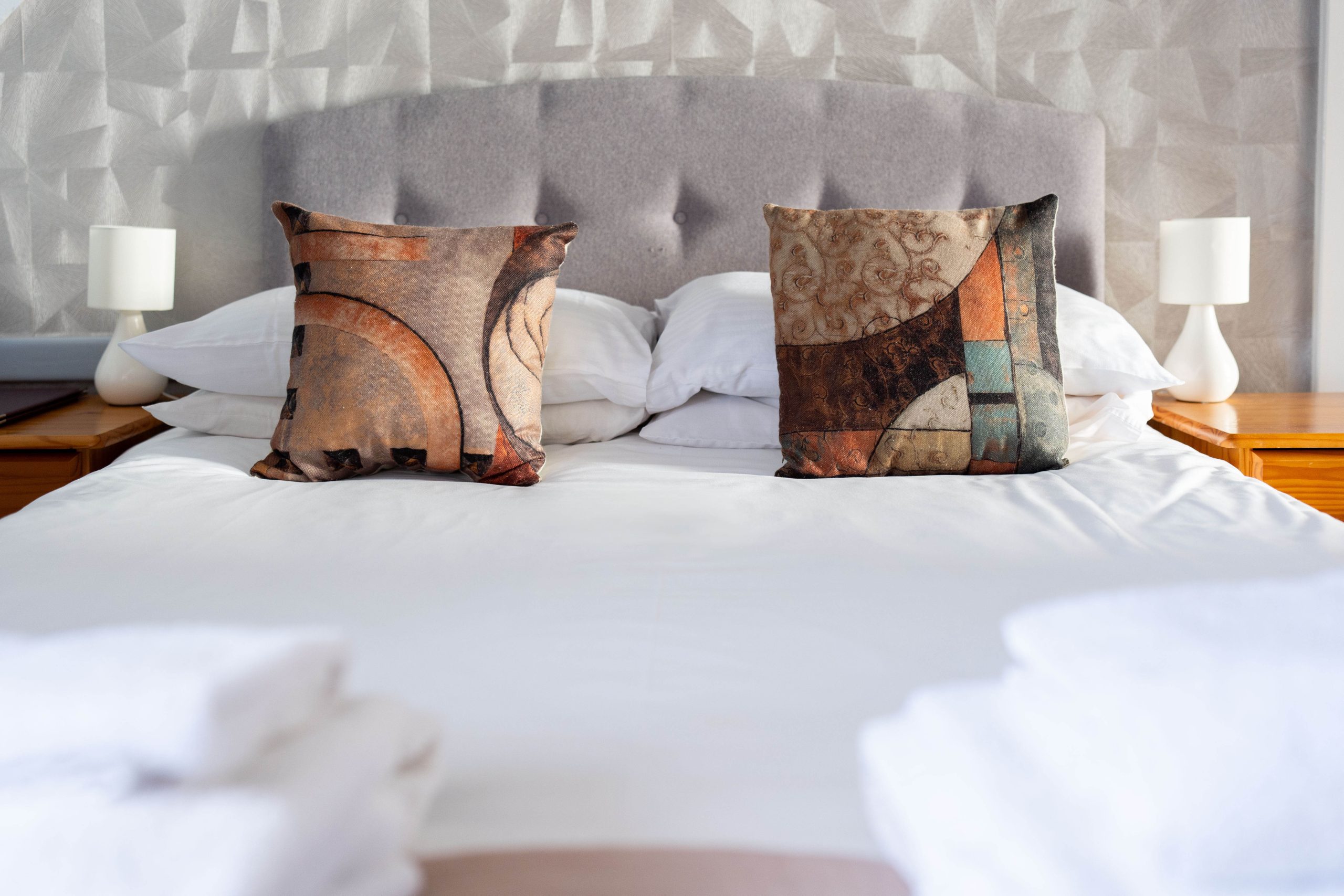 A neatly made double bed with white sheets, two patterned scatter cushions, and a grey upholstered headboard. Wooden bedside tables with white lamps are on either side. Folded white towels are at the foot of the bed.