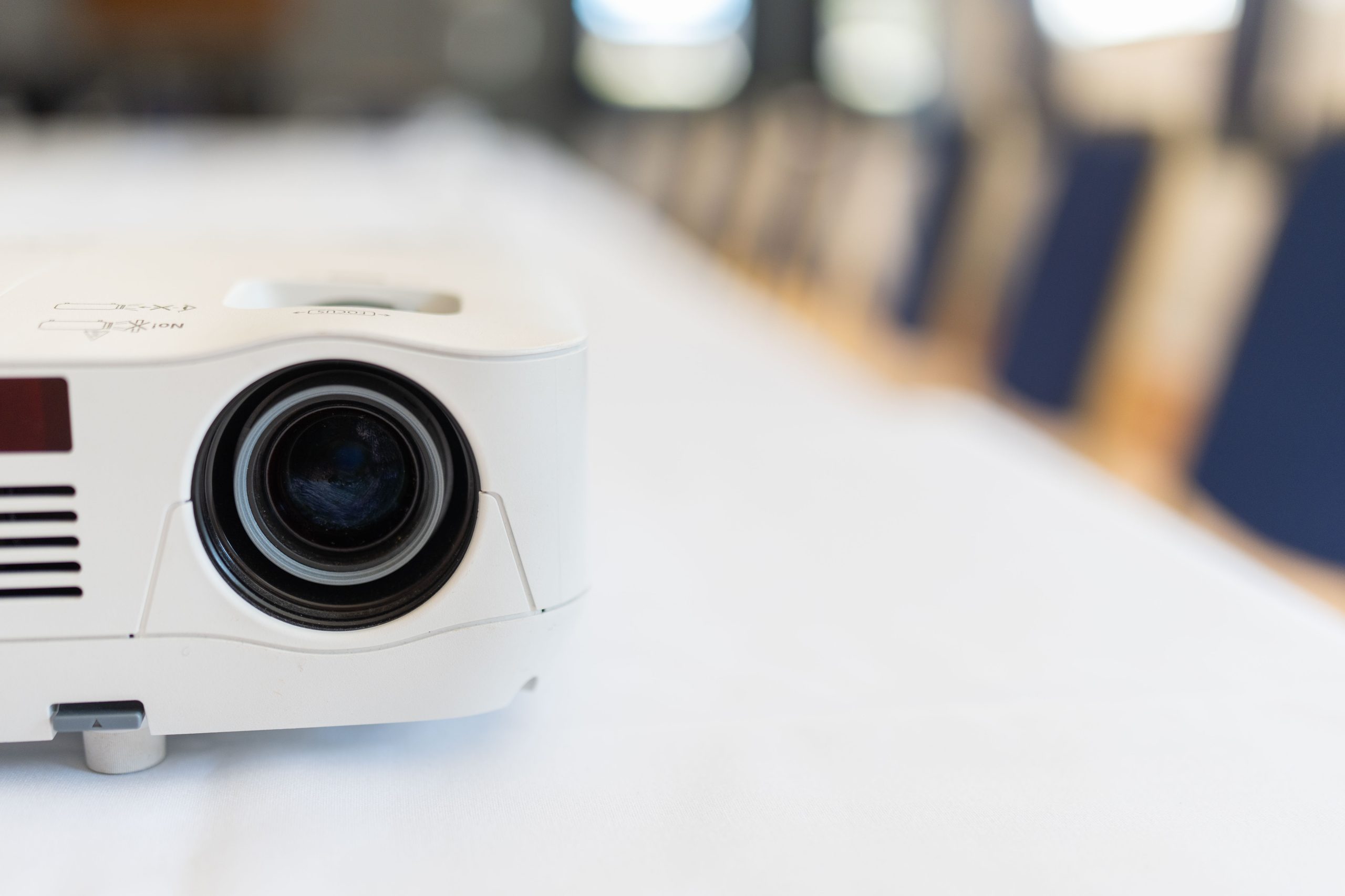 A close-up of a white projector on a table, with rows of empty blue chairs blurred in the background. The focus is on the projector lens, positioned on the left side of the image.