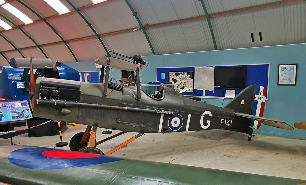 A vintage military biplane with British Royal Air Force roundels and the code F141 on display indoors, surrounded by informational boards and museum exhibits.