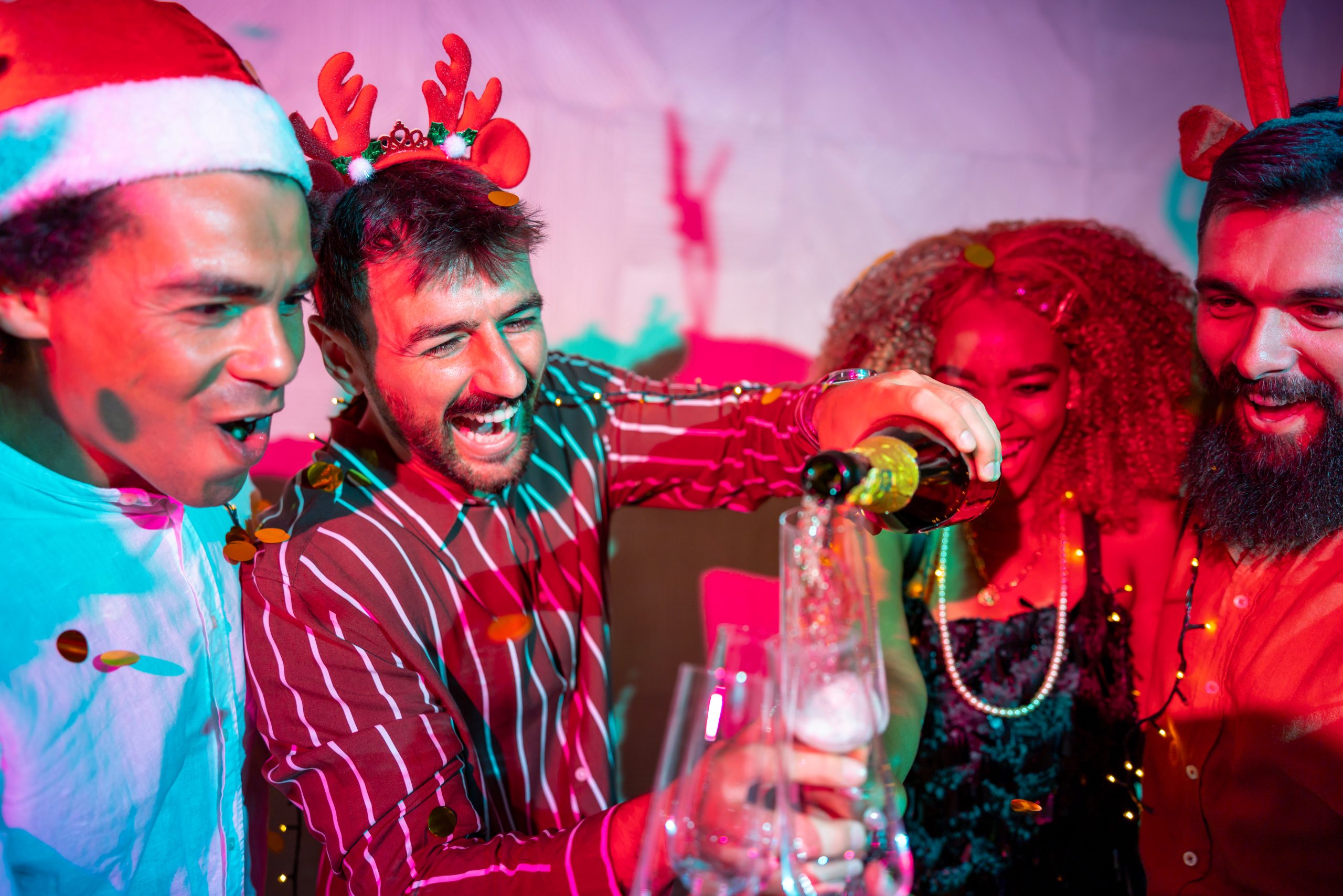 Four adults at a festive party, two wearing holiday headbands, laughing as one person pours champagne into glasses, with confetti and colourful lighting in the background.