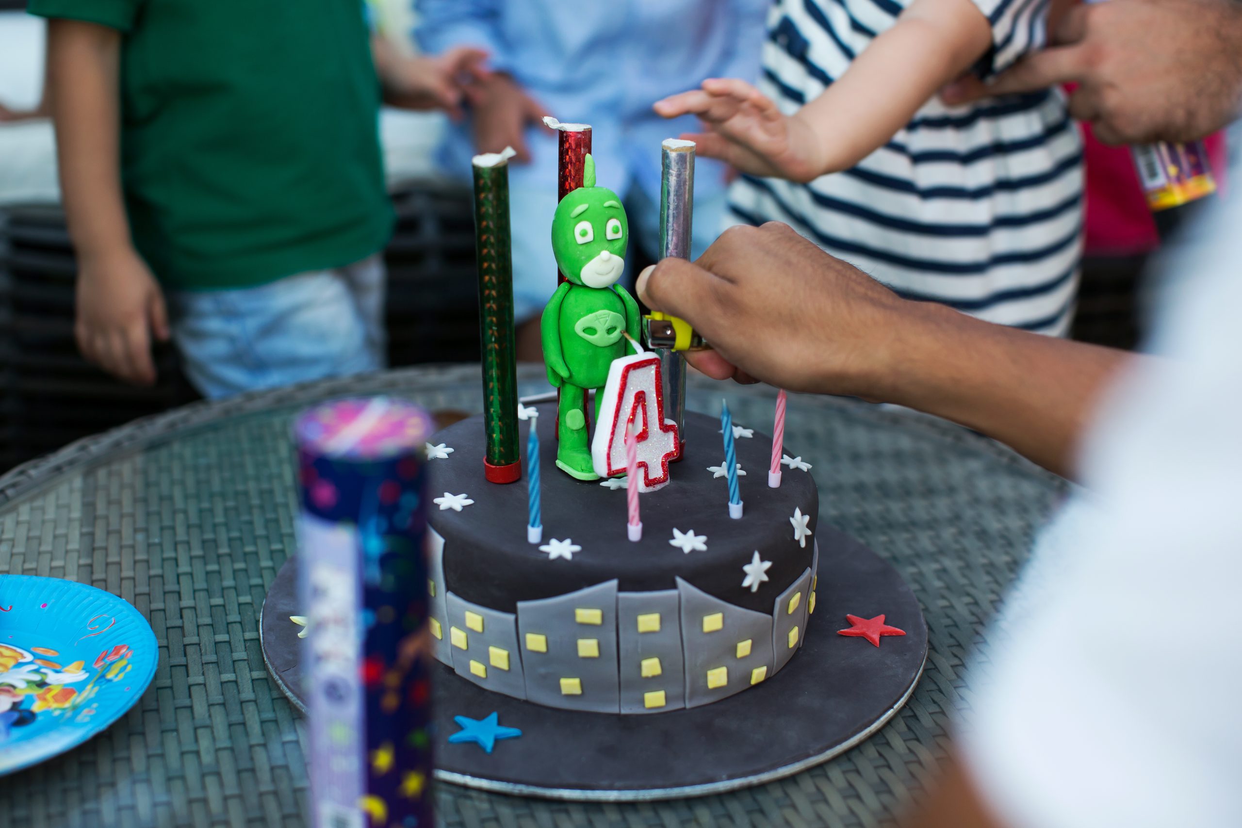 A hand adjusts a number 4 birthday candle on a black cake decorated with cityscape designs and a green cartoon character. Other children and colourful candles are visible around the table.