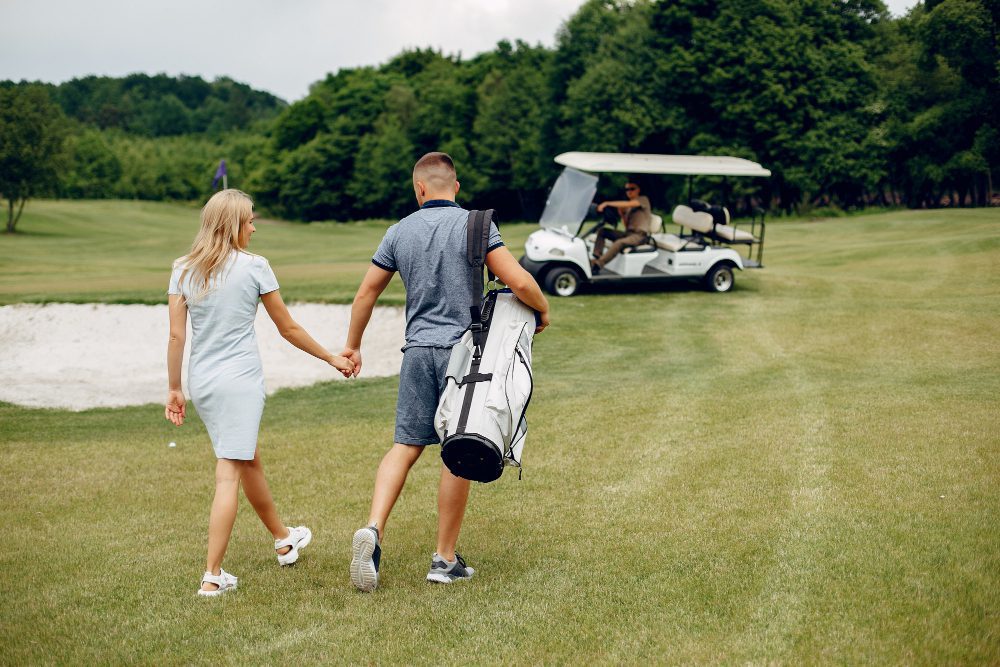 A man carrying a golf bag holds hands with a woman as they walk on a golf course. A golf buggy with two people is visible in the background near green trees.