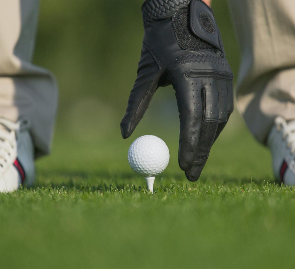 A close-up of a person wearing a black golf glove placing a white golf ball on a tee, with grass and part of their trainers visible in the background.