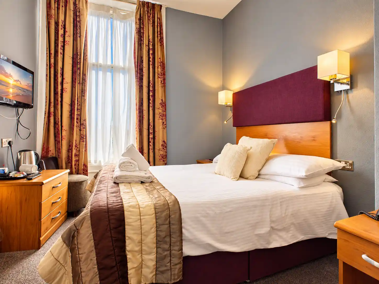 A hotel room featuring a bed with a burgundy headboard, a wooden desk with a television and kettle, and floor-length patterned curtains.