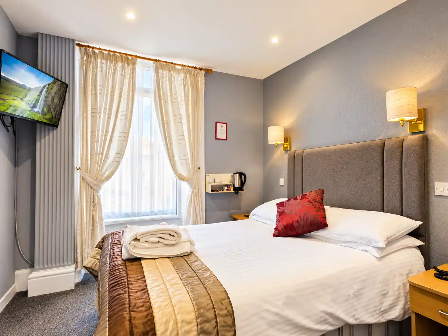 A hotel bedroom featuring a bed with white linen and a brown runner, a television mounted on the wall, and curtains over a window.