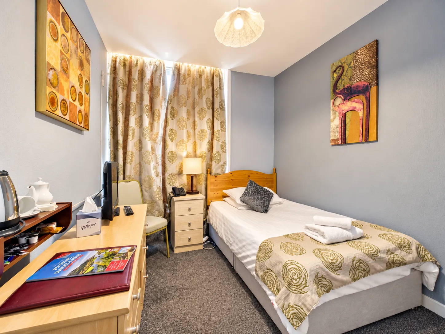 A hotel room with a single bed, a wooden desk with a television, and patterned wall art.