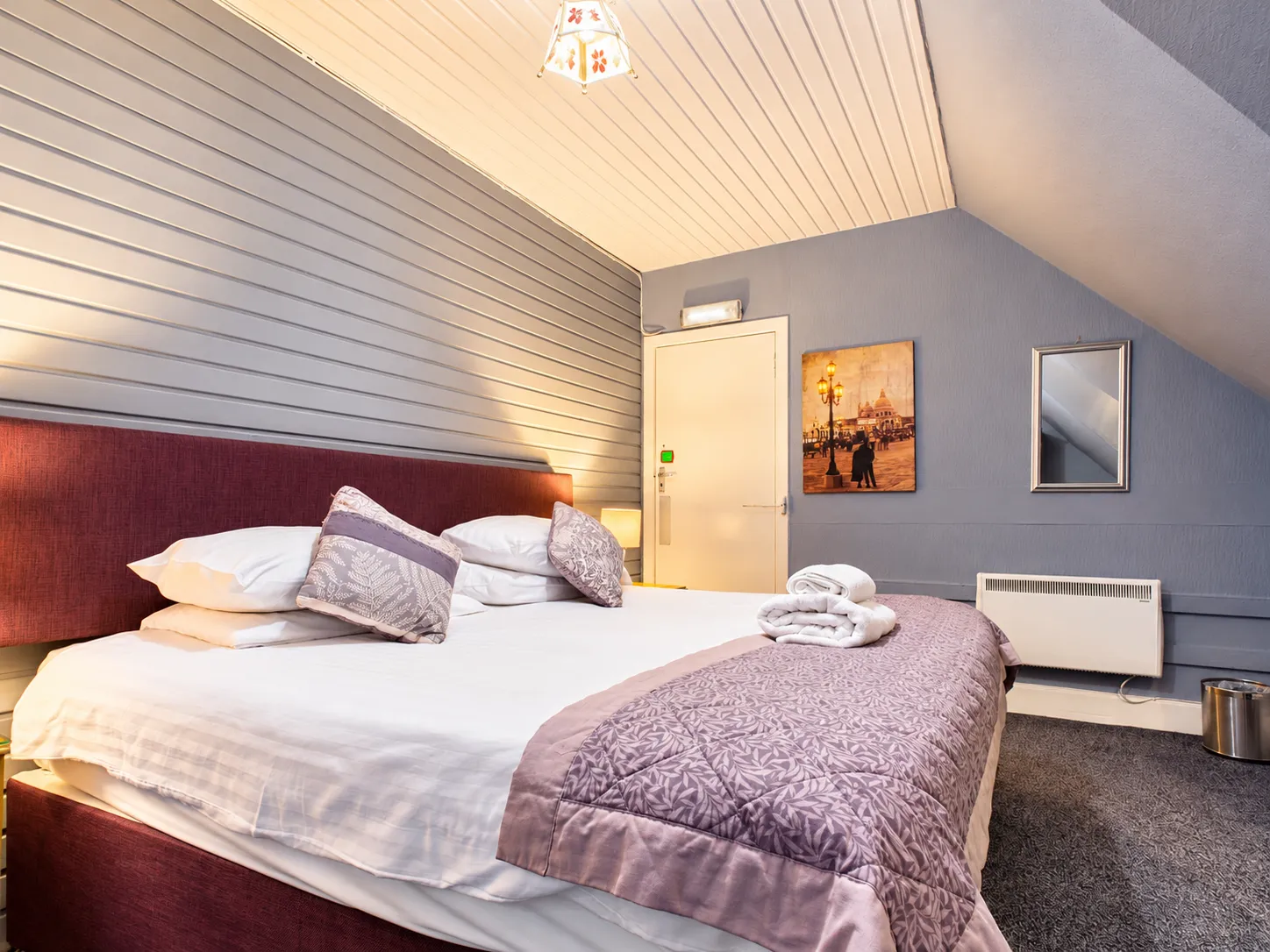 A hotel room with a queen bed featuring a dark red headboard, patterned runner, and folded white towels, with a framed picture on the wall above an electric heater.