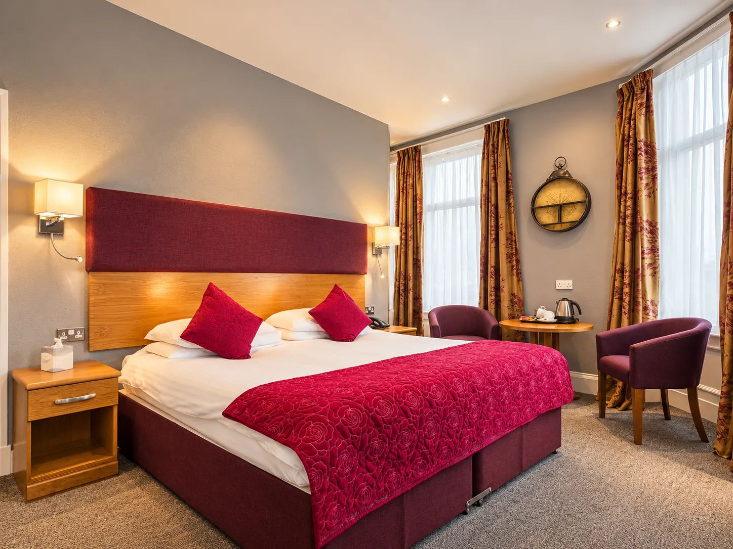 A hotel bedroom with a king bed featuring a deep red headboard and rose-patterned runner, two purple armchairs, and a small wooden table with a kettle.