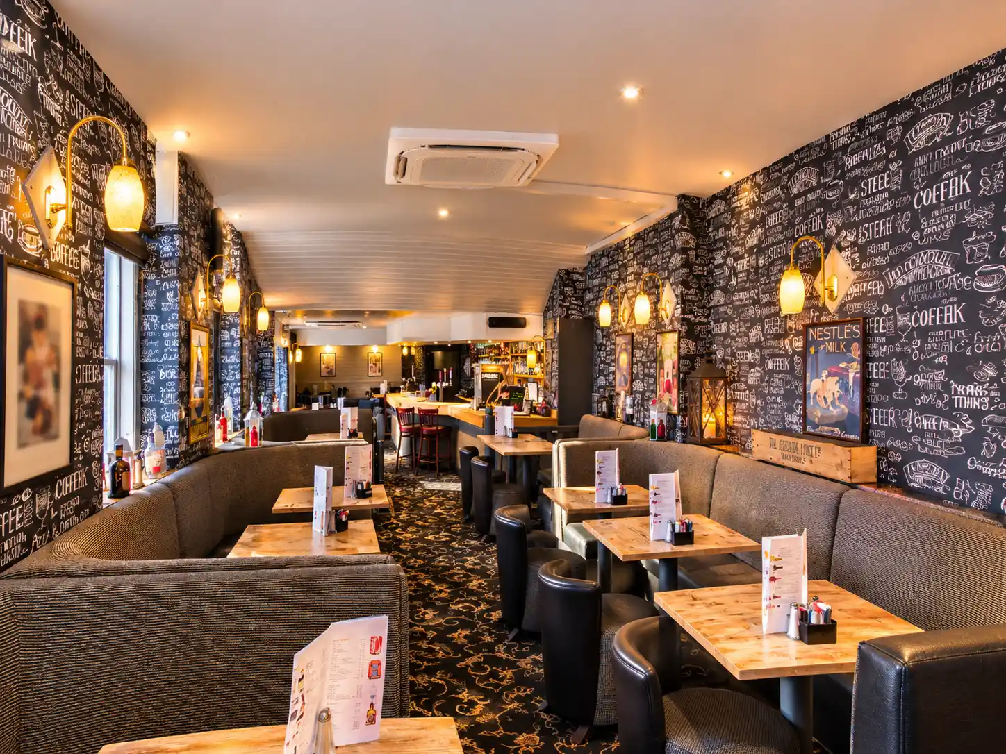 Interior of a pub with rows of wooden tables, grey upholstered booths, and walls covered in coffee-themed chalkboard art.