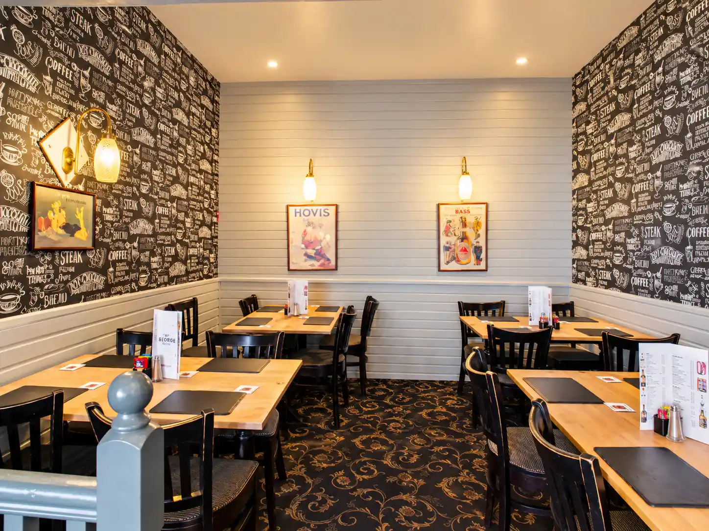 Dining area with light wood tables and dark wooden chairs, featuring black and white typographic wallpaper and two vintage-style posters on a panelled wall.