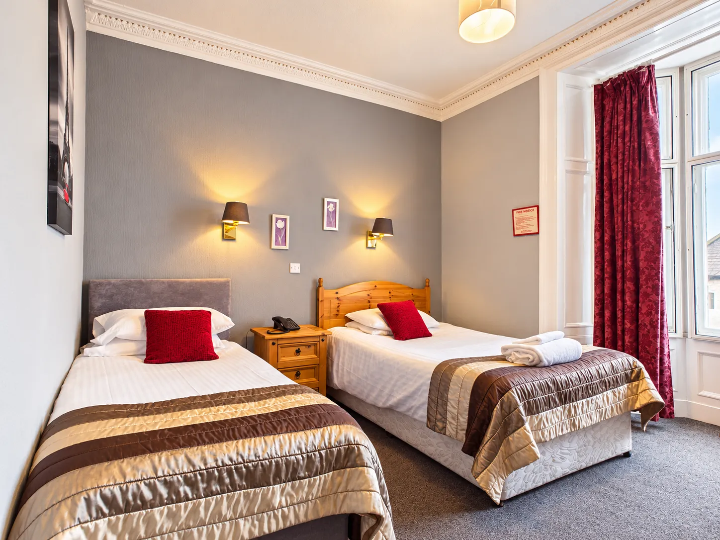 A hotel room with two single beds, each topped with a red scatter cushion and a brown quilted runner.